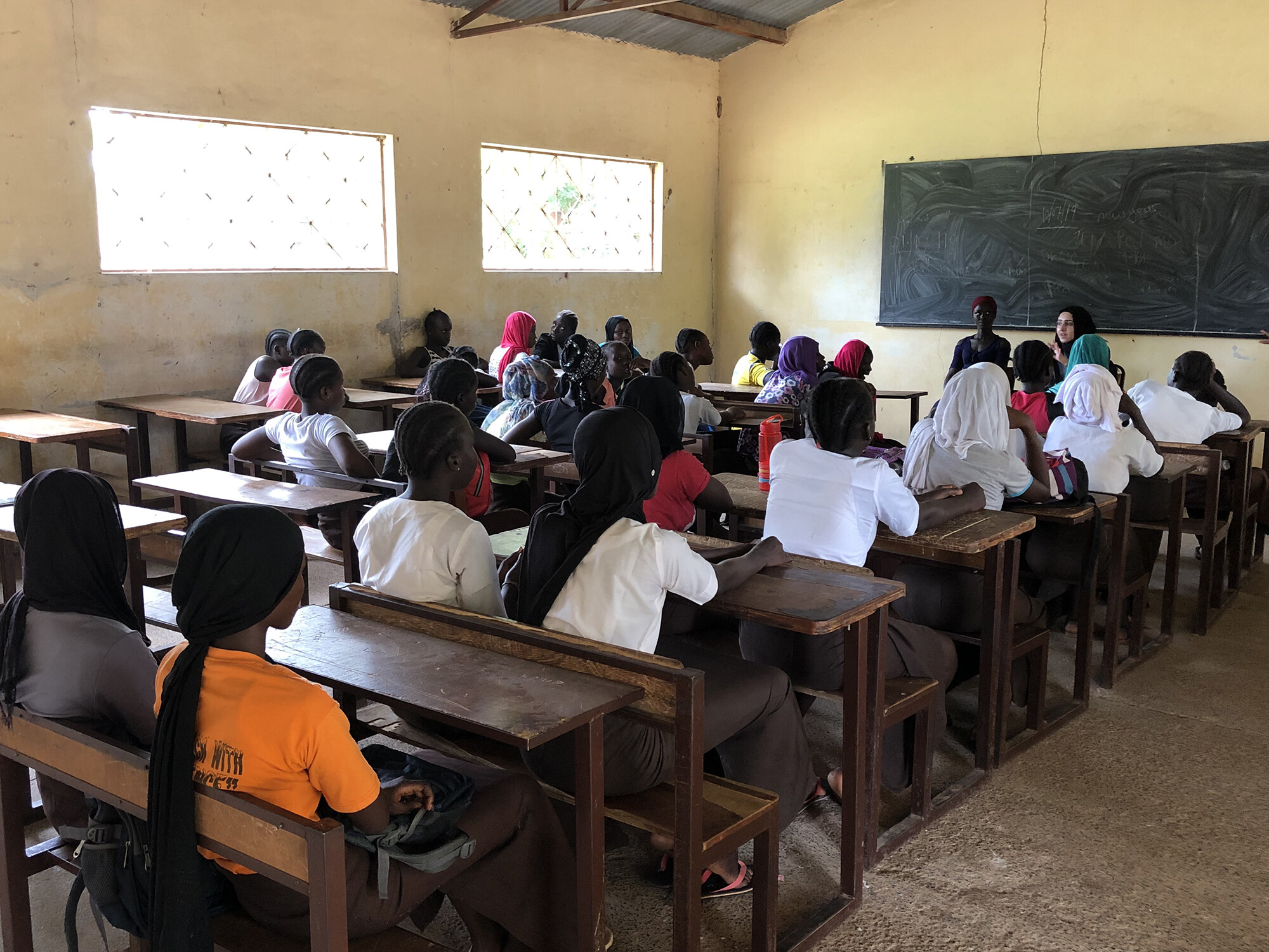 classroom full of girls seated at wooden desks, Fatima Al Rashed is seated at the front of the room in front of a blackboard.