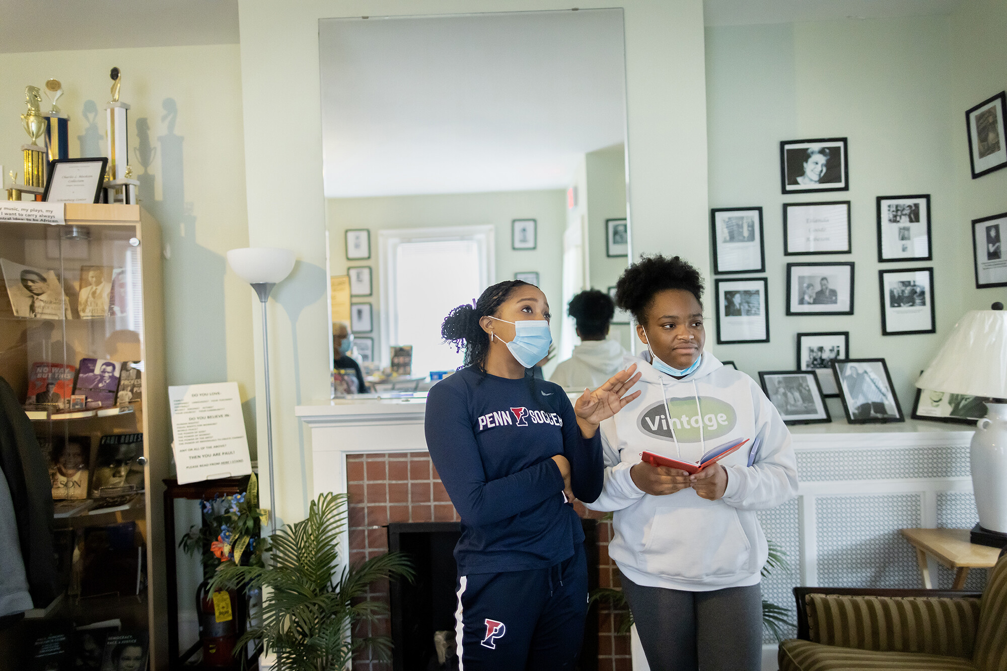Two woman converse in a house museum with images and trophies placed on shelves around a mantle.
