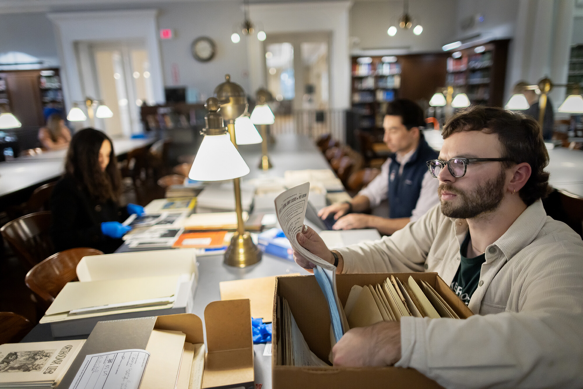 Sam Herrmann looks through a banker box full of files at a long table in a reading room. 