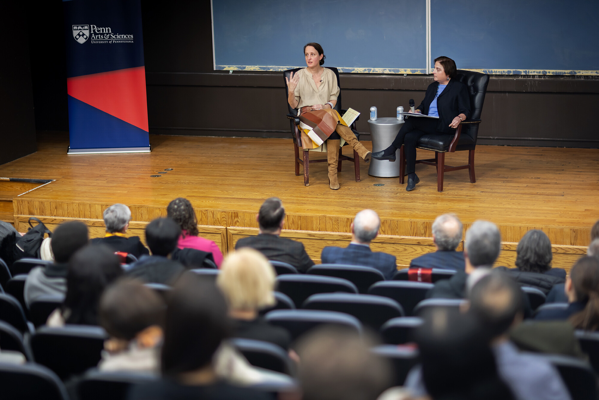 Sophia Rosenfeld and Beth Wenger sit on chairs on a wooden stage in front of a black board and a sign reading Penn Arts & Sciences, as Rosenfeld addresses the audience.
