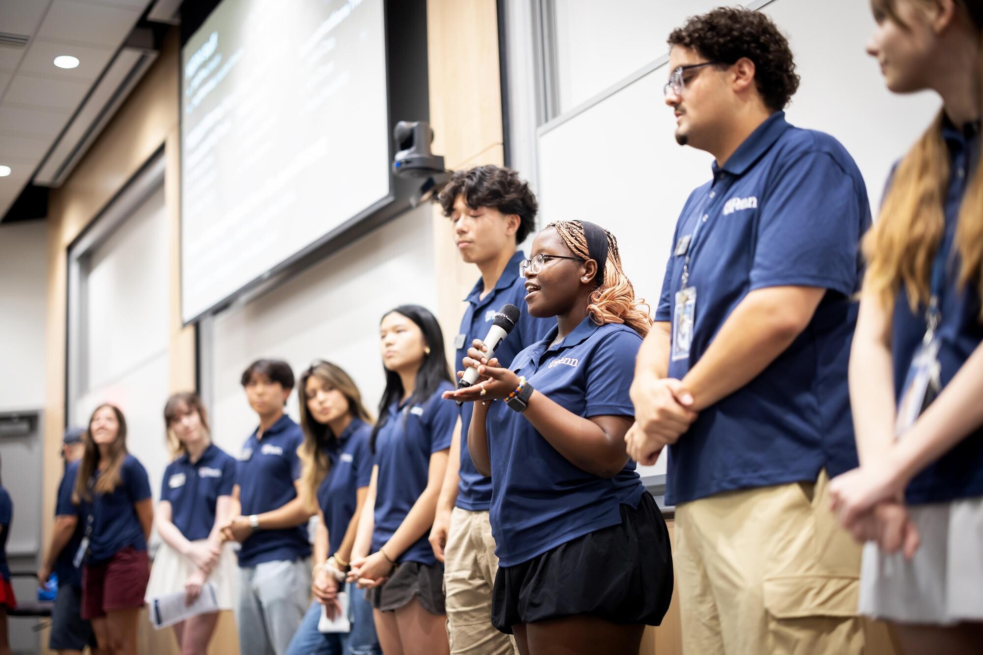 Student volunteers lined up, one is addressing the room while holding a mic.