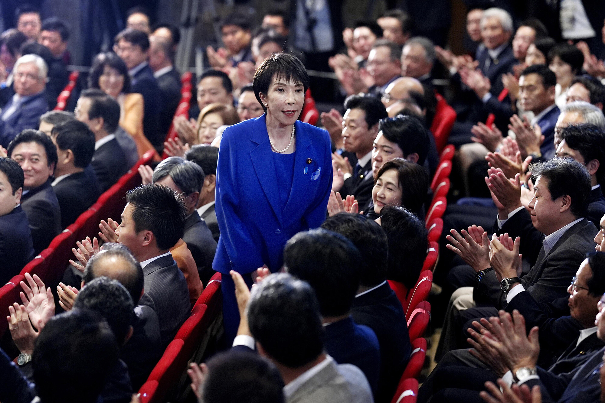 Sanae Takaichi, wearing a blue suit, stands in a crowd of seated Japanese legislators.