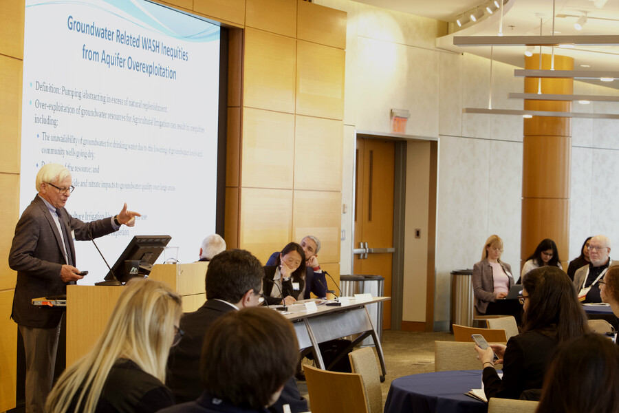A speaker gestures at a podium, as panelists and audience members listen. His slide reads "Groundwater Related WASH Inequities from Aquifer Overexploitation"