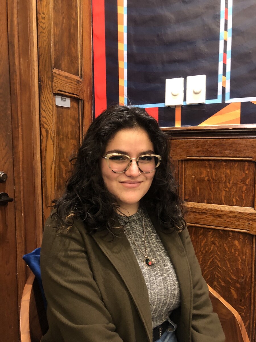 Person wearing glasses smiles at the camera with a wooden door and wall behind herZu
