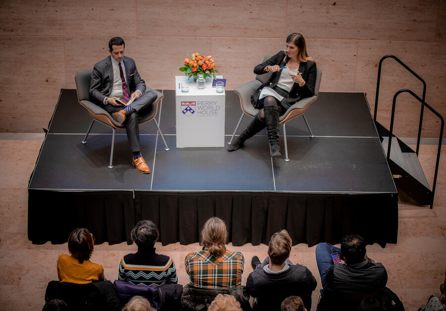 Two people sit in chairs in a discussion, with a table between them bearing a sign reading Perry World House, with a vase of flowers and glasses of water on top