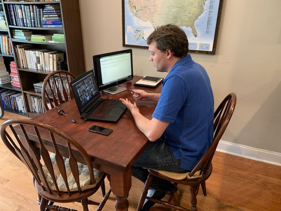 Person sits at wooden kitchen table looking at two computers, with a map of the United States on the wall behind him.
