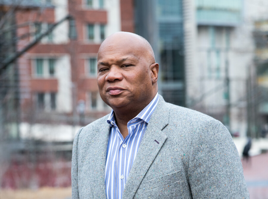 A person standing outside, brick buildings blurry in the background, wearing a gray wool blazer and a blue-and-white-striped button down shirt.