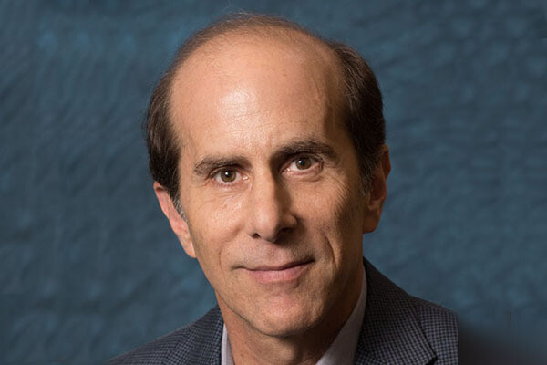 Close-up headshot of a person wearing a tweed blazer and a purple-gray button down shirt in front of a blue background.