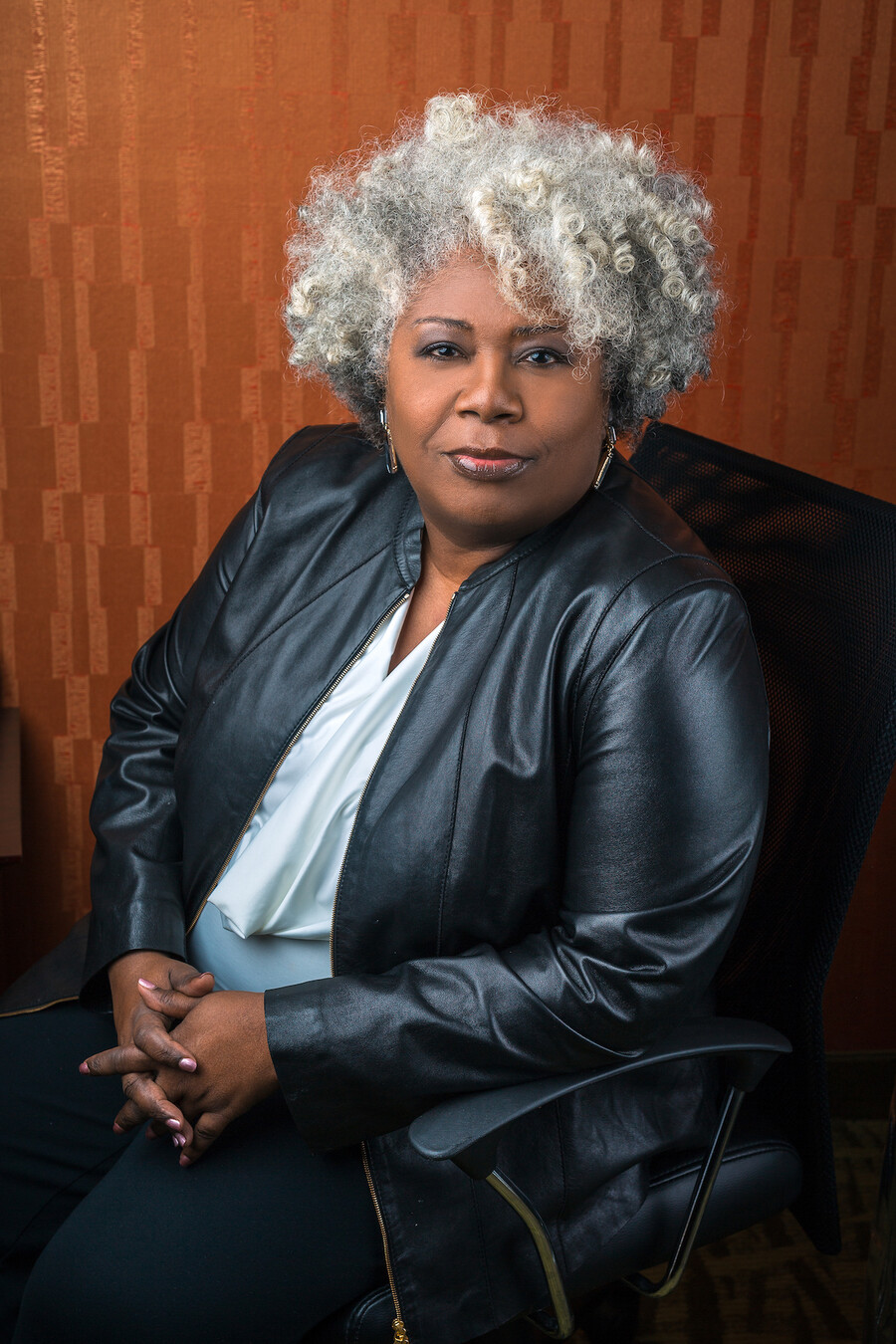 Woman in dressy black leather jacket, silk grey blouse and silver earring sits in a black chair and looks at the camera, hands folded 