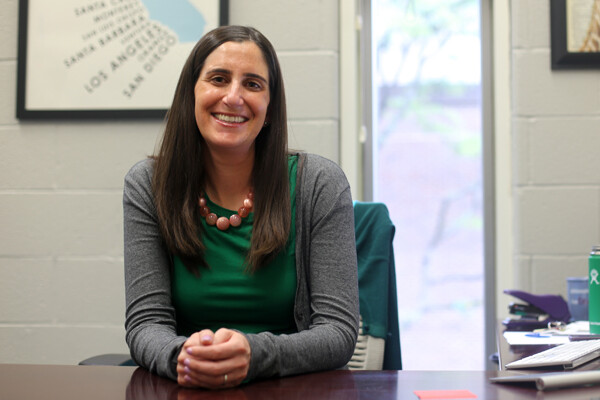 Woman in gray cardigan, green shirt and brown beaded necklace sits at a desk with hands folded, smiling at the camera, a white painted block wall behind her and a tall, slim window showing trees outside.