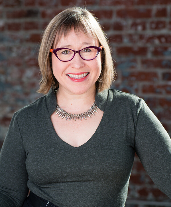 Woman with glasses, a silver necklance and grey shirt stands in front of a brick wall, smiling at the camera