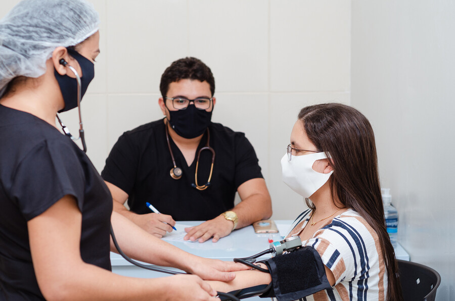 Two masked medical professionals attend to a masked patient, taking their blood pressure.
