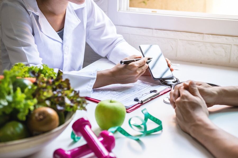 doctor and patient seated at a desk with a medical chart, smartphone, bowl of vegetables, hand weights and measuring tape.