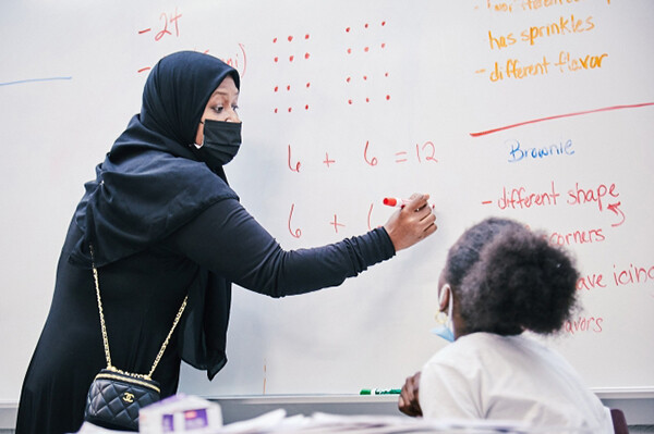 A teacher writing math problems on a white board in front of an elementary school student.