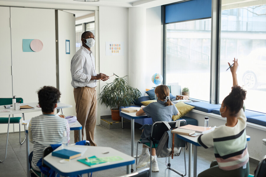 Masked teacher calling on a young student raising their hand in a classroom.