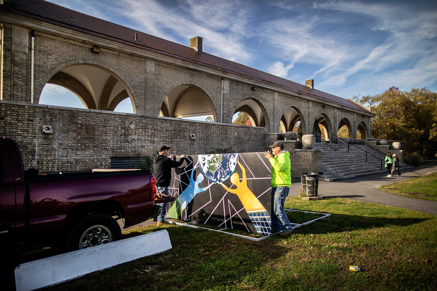 Two people holding a large painting that will be mounted on a wall of an outdoor portico.