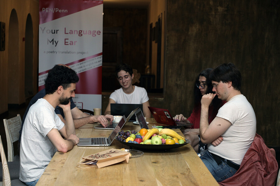 People sit around a table with a bowl of fresh fruit in front of a sign reading Your language my ear.
