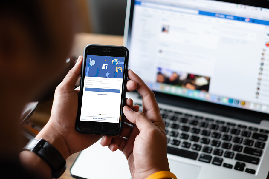 Hands holding a smartphone open to Facebook in front of a laptop open to Facebook.