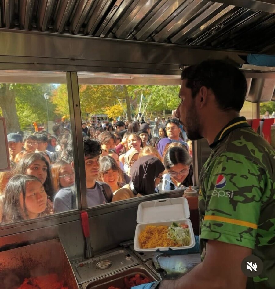 Rahim Ullah prepares food for a student from inside his food truck. Outside the truck window can be seen a crowd of students.