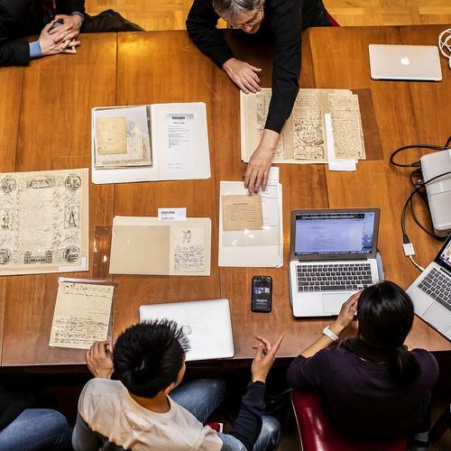 Overhead shot of rare documents spread out on wood table with students and professors and laptops surrounding them. 