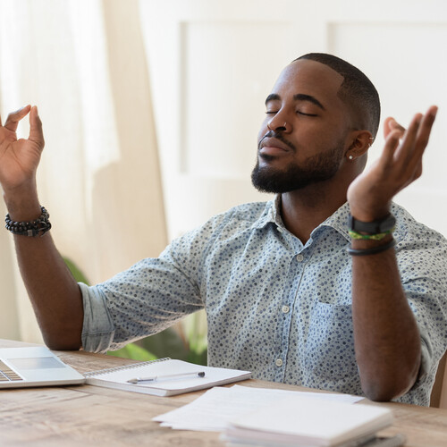 person in sunlit room with laptop and notebook with eyes closed and arms open having a meditative moment.