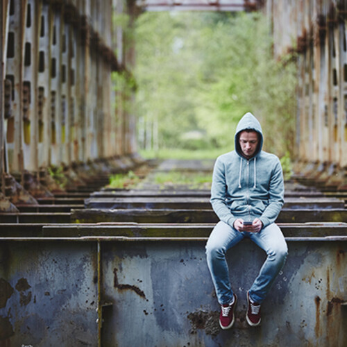 Person wearing a hoodie sits on a structural beam alone in an isolated area holding a smartphone.