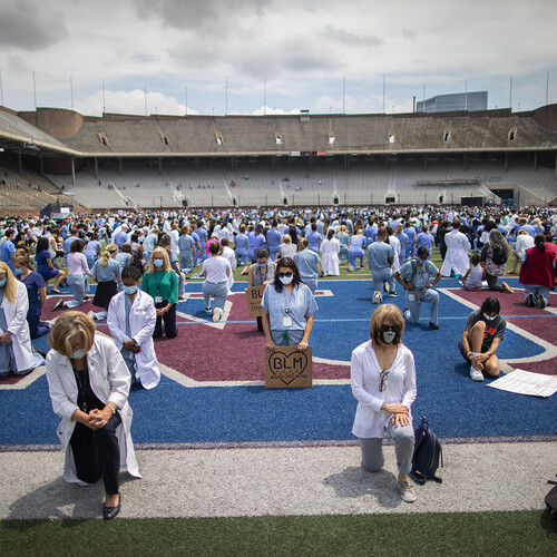 Medical personnel in scrubs, white coats and face masks kneel on the field at Franklin Field, one holds a sign with the letters BLM surrounded by a heart.