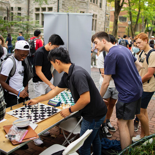 students playing chess outside on a table with several others watching