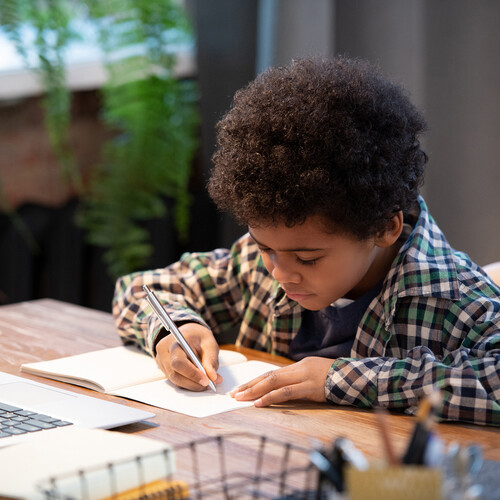 Young child writing in a notebook at a table with a laptop.