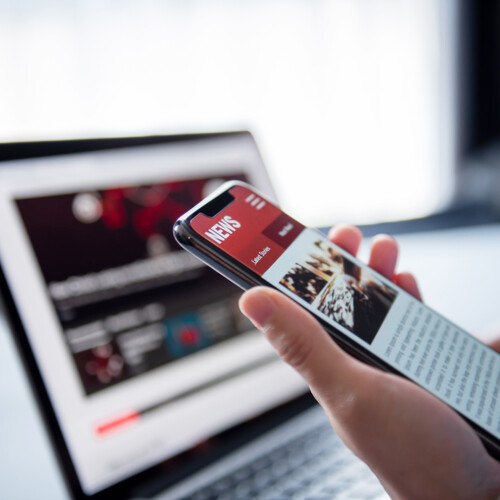 A hand holding a smartphone with news info in front of a laptop open to news.