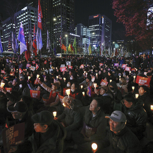 A crowd of people are gathered in a public square, facing left, holding candles and carrying signs in a vigil calling for the South Korean president’s resignation.