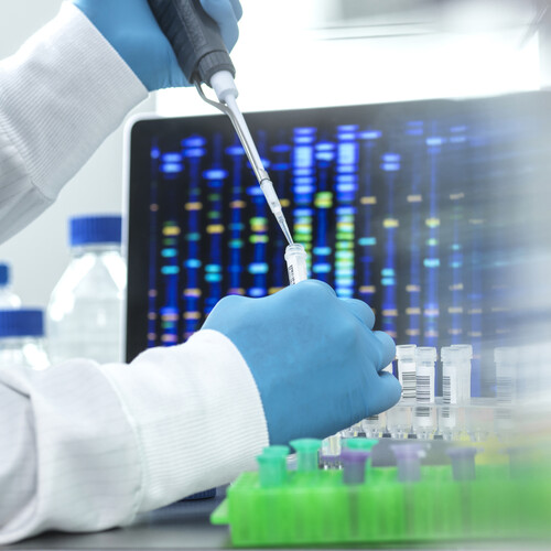 A scientist using a pipette in a lab with a computer screen of DNA sequences on it.