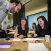man leaning over table where two seated women are working with art tools on paper