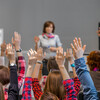 Seated students in a classroom raise their hands while a teacher stands at the head of the classroom