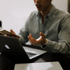Person sitting at personal laptop holding hand out as though in discussion virtually.