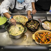 The arms of two people over an industrial-sized kitchen warmer, which holds a pot of green puree, two pans of pasta, a cast iron skillet of mushrooms, another cast iron skillet of fish with a spatula on top, and a bowl of multi-colored carrots. A stack of bowls sits off to the left of the image.