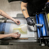 two sets of arms over a hand-operated printing press, one set with gloved hands putting ink on a metal cylinder and the other placing a printing plate with an image of a tree without leaves on the flat surface in front of the cylinder