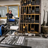 two people working the hand-operated press in the background with wood and metal letters in a shelf in the foreground