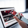 A hand holding a smartphone with news info in front of a laptop open to news.