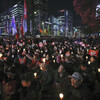A crowd of people are gathered in a public square, facing left, holding candles and carrying signs in a vigil calling for the South Korean president’s resignation.