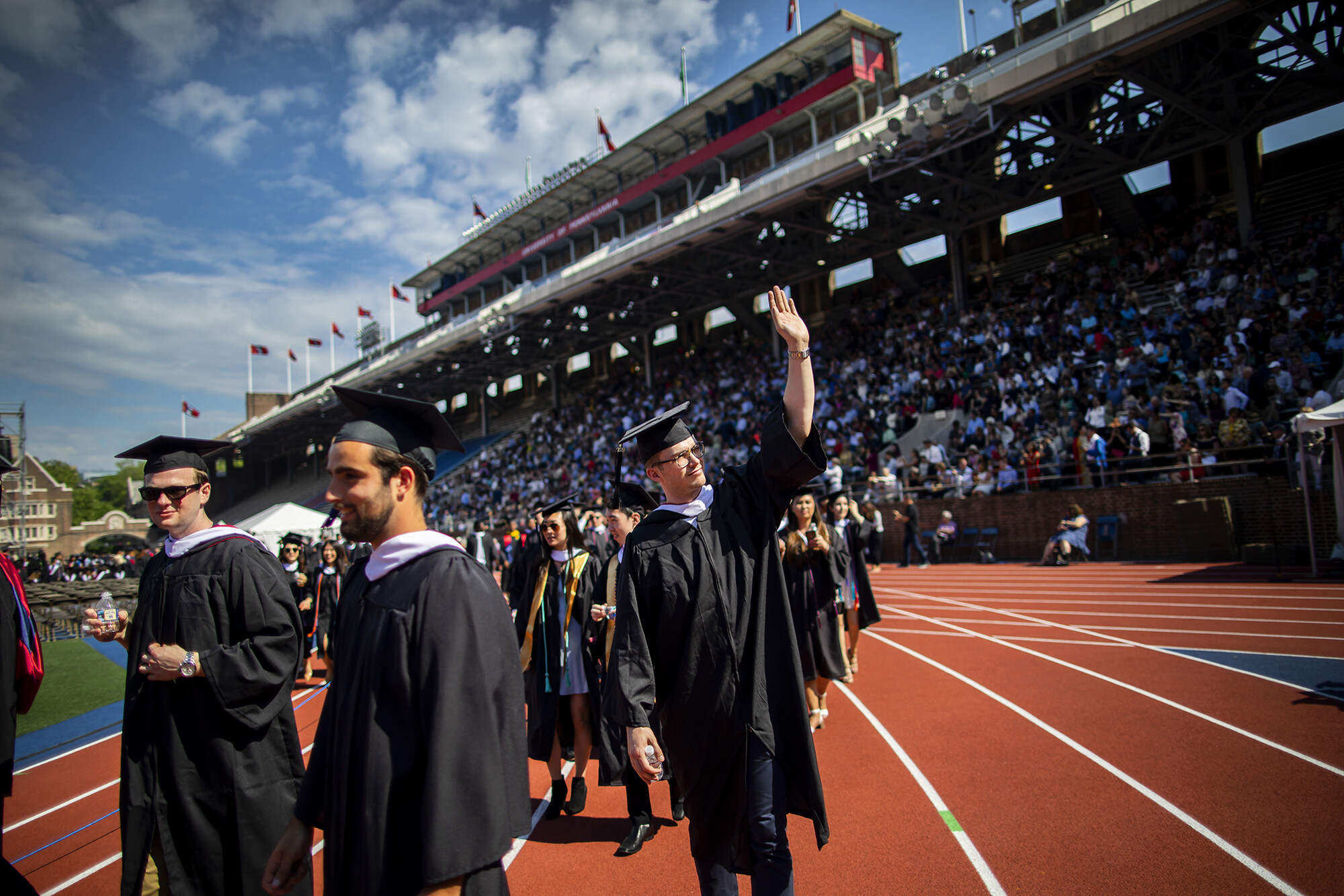 Graduates arrive on Franklin Field, one waving to audience.