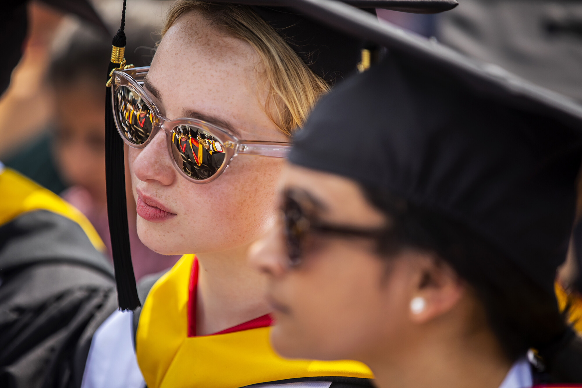 Graduate in sunglasses seated with reflection of other graduates in sunglasses.