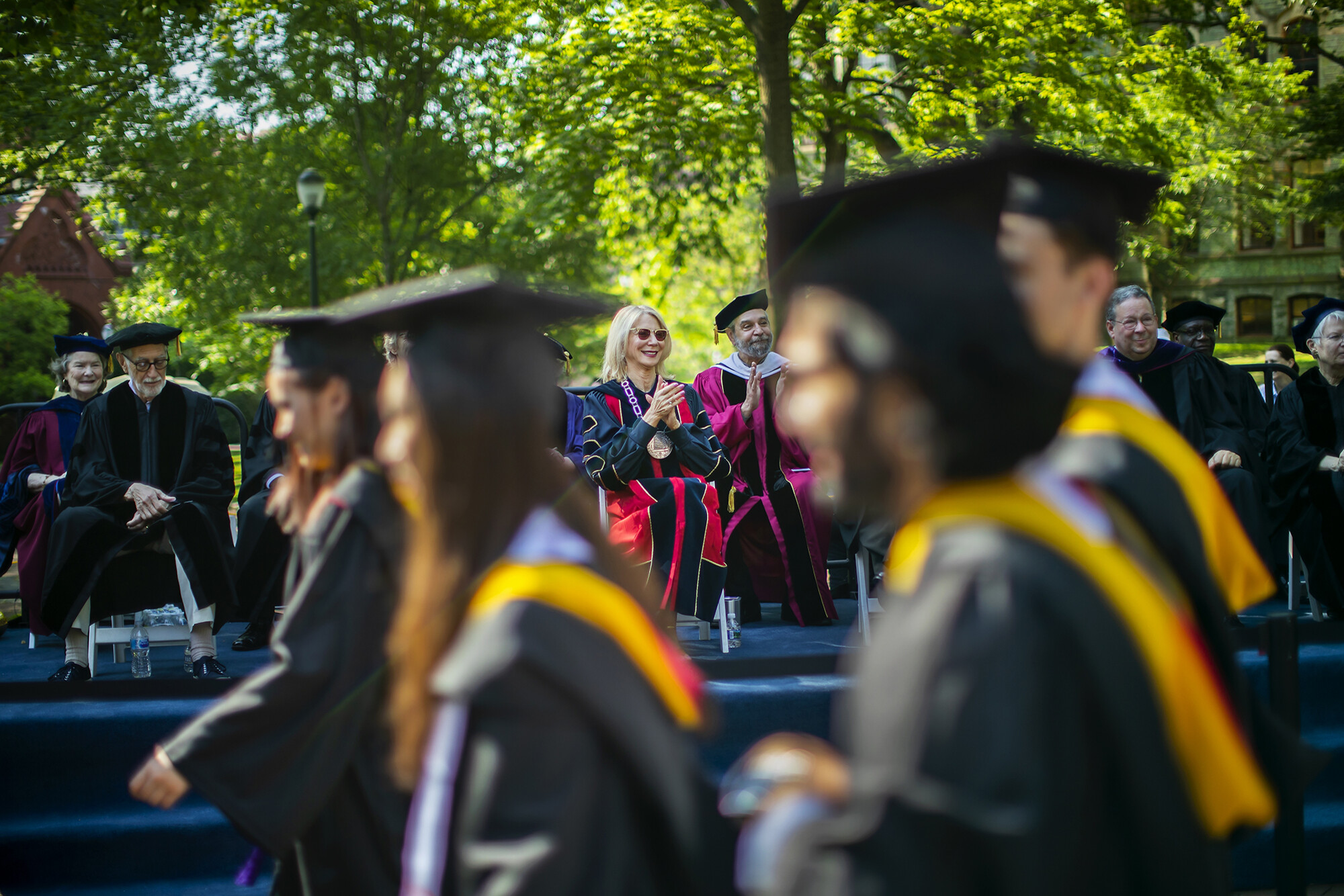 Amy Gutmann and Michael Delli Carpini are seated and clapping as procession of graduates walk past small risers on Locust Walk.
