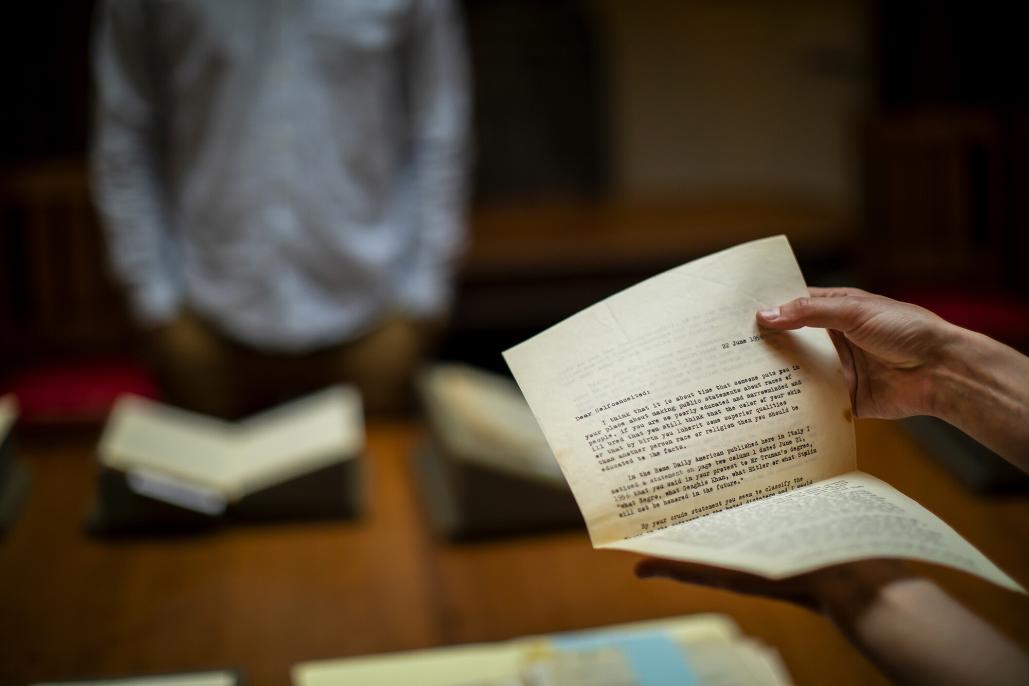 Typed letter, many pages thick, being held by two hands. A blurred figured standing in front of a book is in the background.