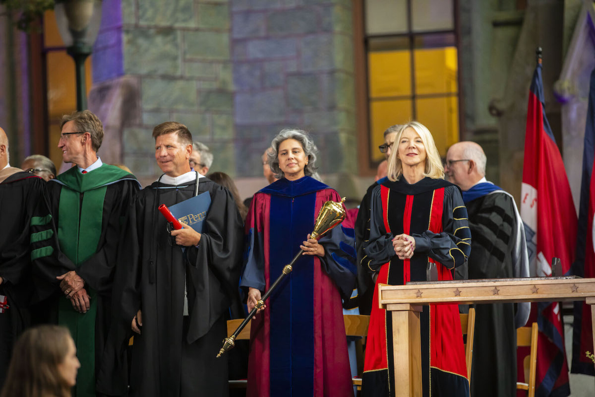 Several people in academic regalia onstage in front of College Hall, including Amy Gutmann at left and Medha Narvekar to her right, holding a mace. 