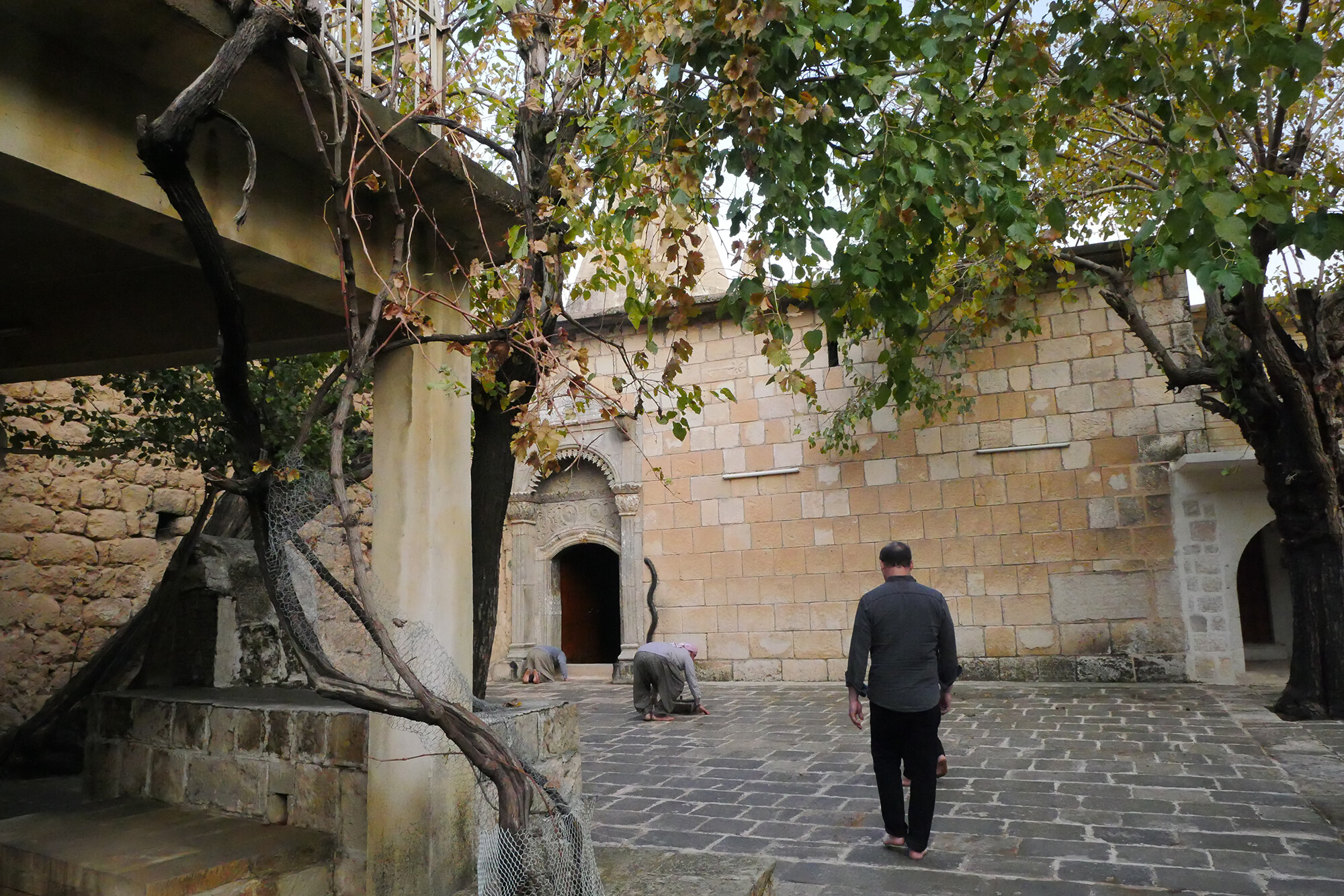 Two people in an outside courtyard at the Lalish Temple in daylight under a tree