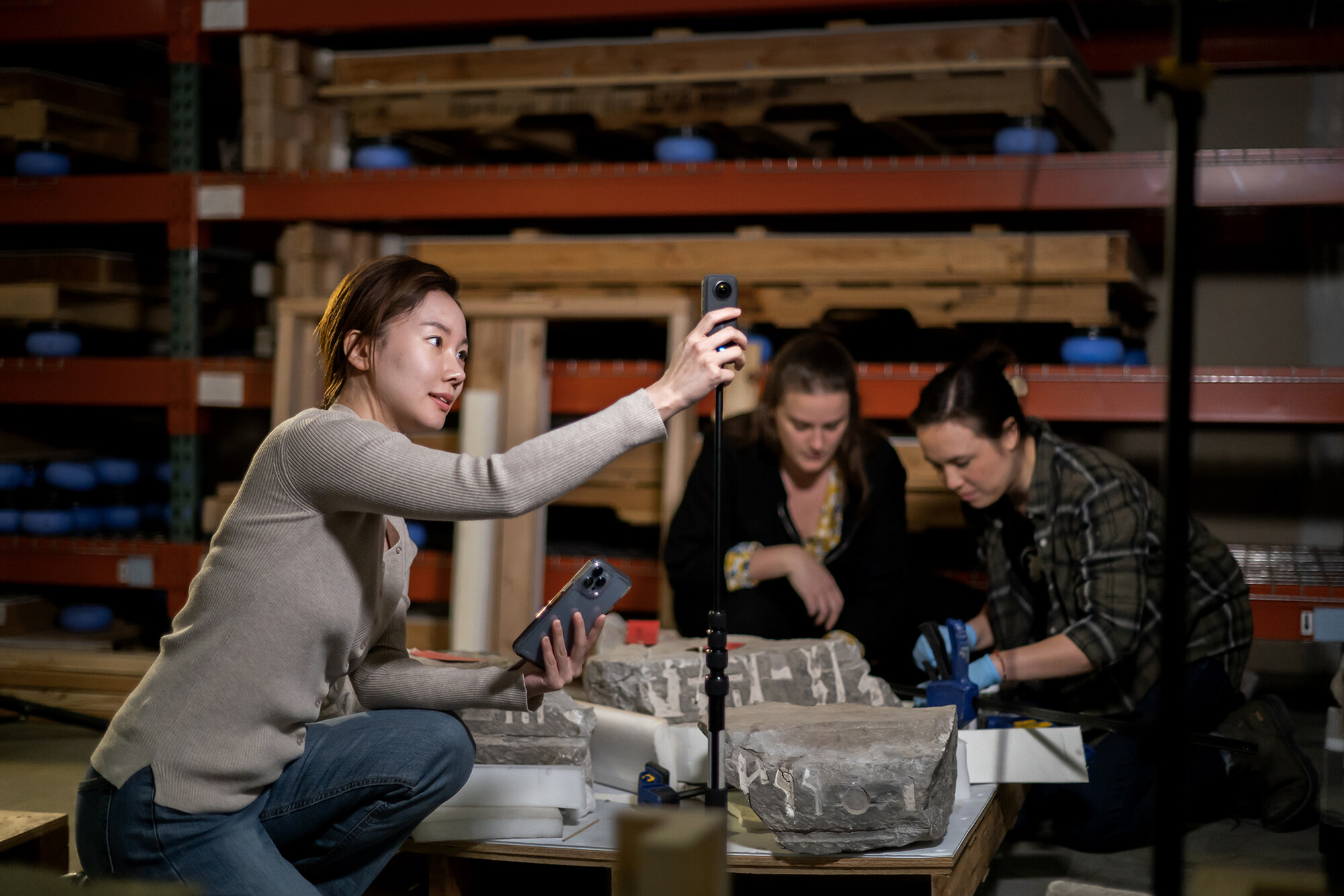 A student checks a light meter while two other students work on the ground of the Museum warehouse.