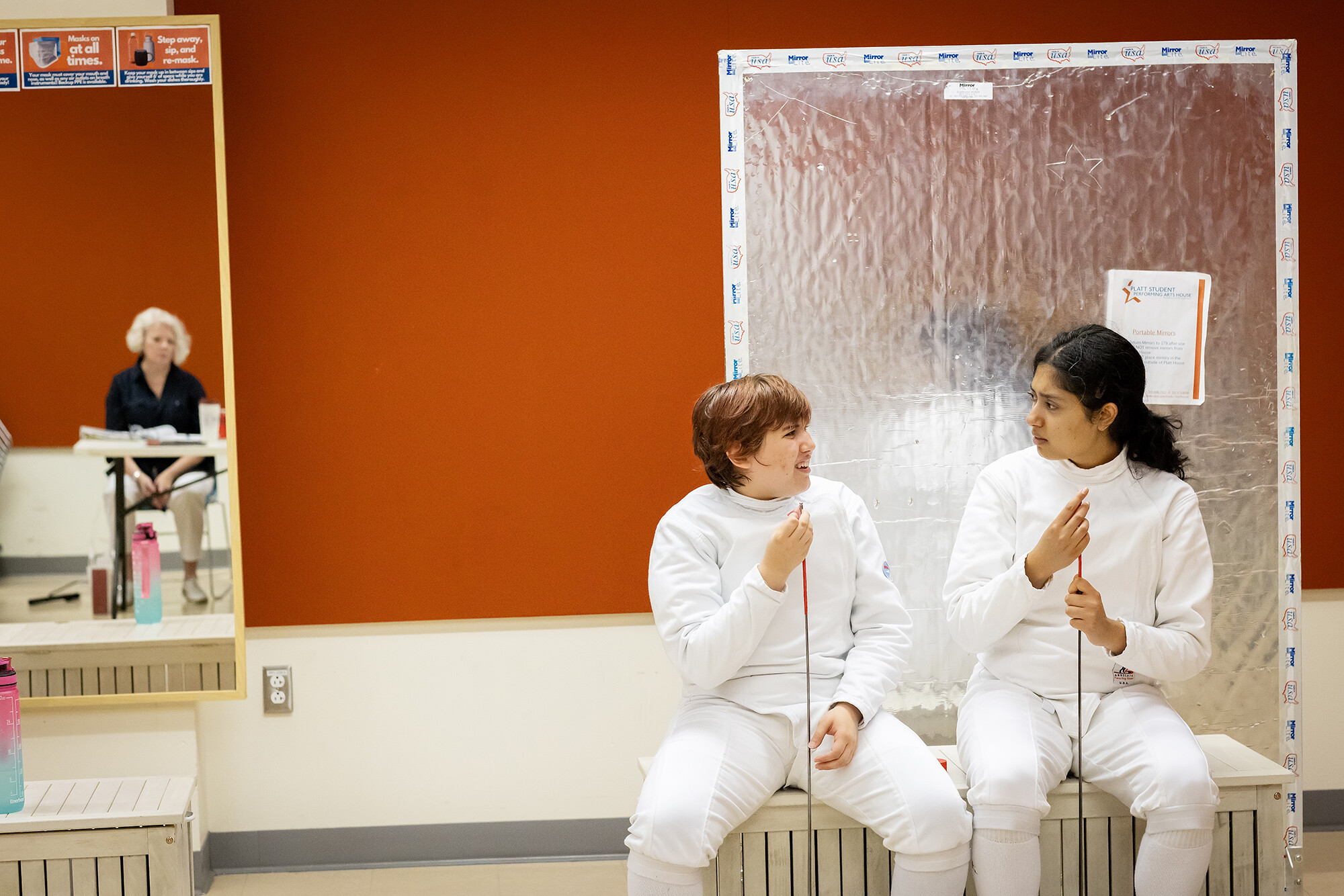 Two actors seated during play rehearsal  in fencing gear with a director’s reflection in a mirror.