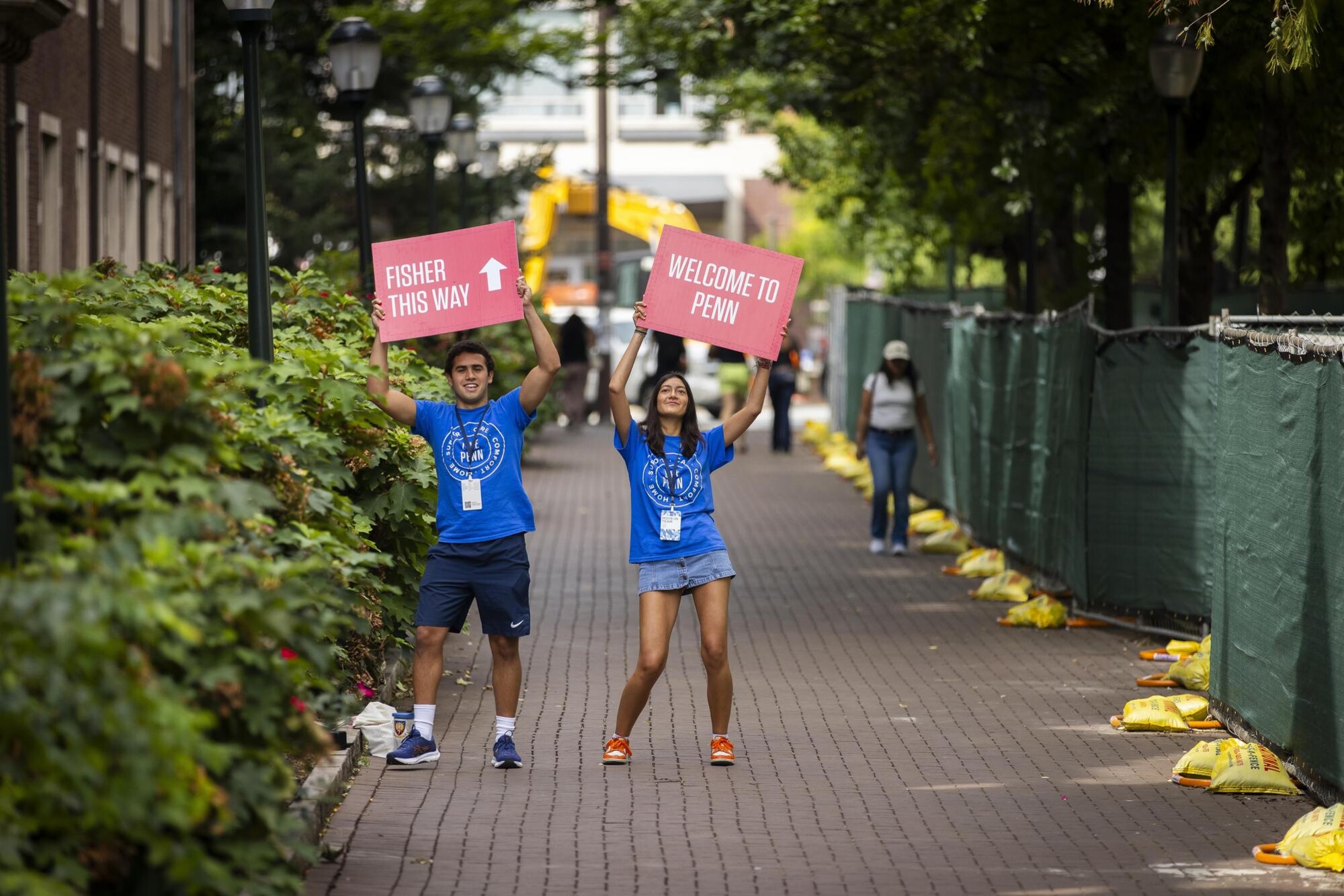 Two Move-In coordinators hold up signs that read Fisher This way and Welcome to Penn on Penn’s campus.