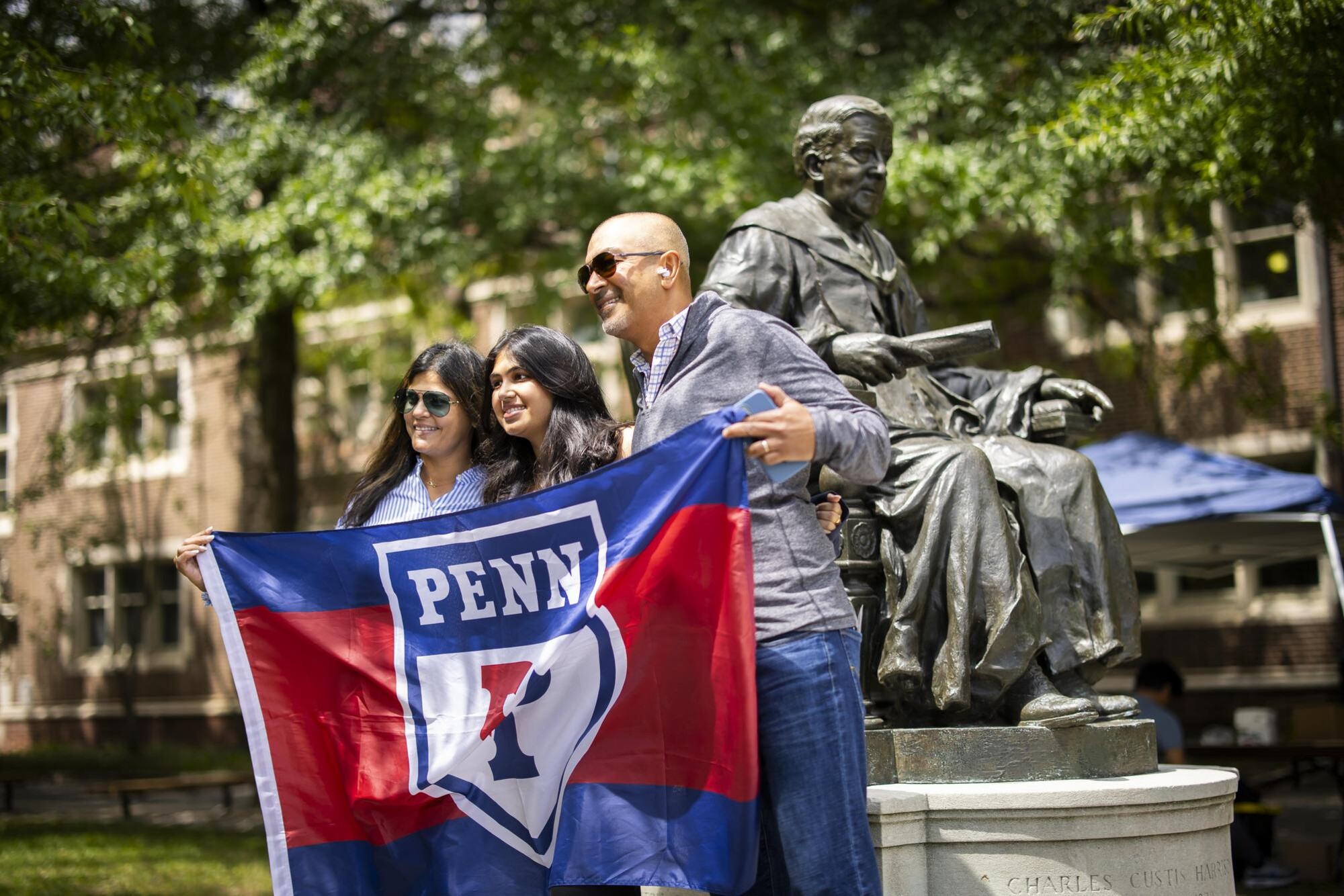 A Penn student and their parents hold up a Penn banner in front of a statue on campus during Move-In.